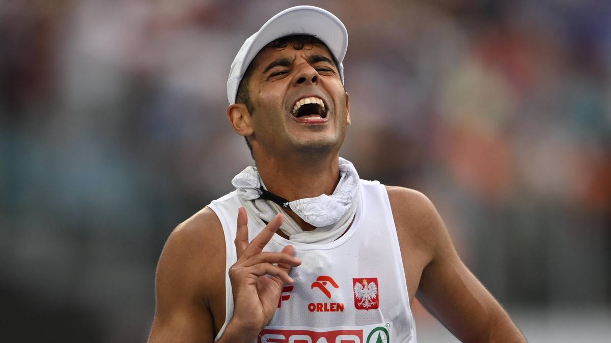 Lekkoatleta w białej koszulce i czapce z daszkiem, z otwartymi ustami i przymkniętymi oczami, wykonuje gest dwoma palcami, biegnąc.