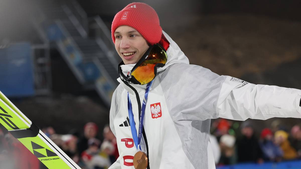 Portret uśmiechniętego Kacpra Tomasiaka w stroju reprezentacyjnym z medalem na szyi i nartami w dłoni.