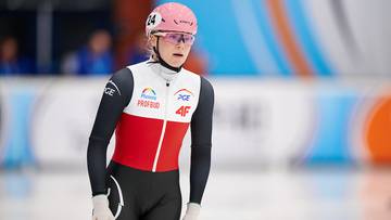 Łyżwiarka szybka w różowym kasku i okularach, w biało-czerwonym stroju sportowym, stoi na lodowisku.