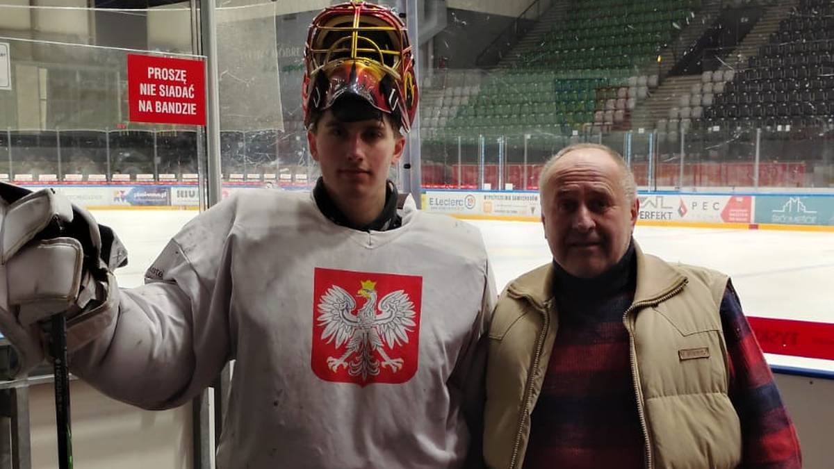 Młody bramkarz hokejowy w stroju z polskim godłem stoi obok starszego mężczyzny na tle lodowiska.
