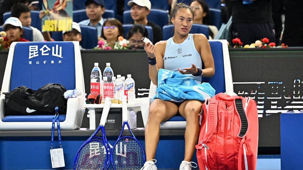 Tenisistka siedzi na krześle z rakietami tenisowymi, butelkami wody i torbą obok. Jest skupiona i trzyma coś w dłoni.