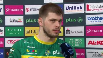 Młody mężczyzna w zielonej koszulce z ż&oacute;łtym kołnierzykiem, z kr&oacute;tkimi jasnymi włosami i zarostem, patrzy w bok, trzymając mikrofon z logo "Polsat Sport". W tle widoczne są kolorowe panele z logotypami sponsor&oacute;w.