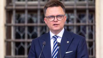 Mężczyzna w eleganckim granatowym garniturze i krawacie, w okularach, przemawia za mównicą z dwoma mikrofonami.