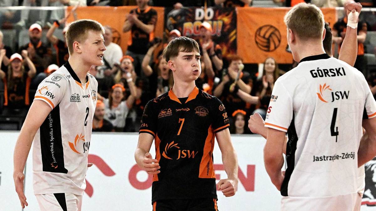 Trzech młodych siatkarzy w strojach sportowych na boisku podczas meczu, z kibicami na trybunach w tle.