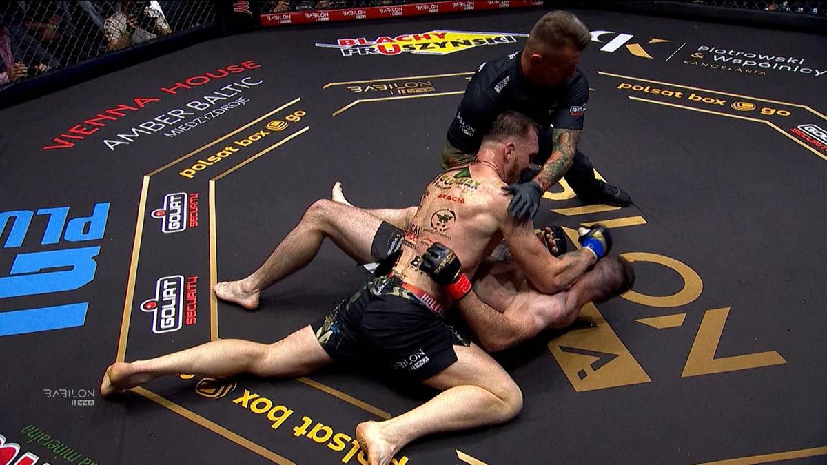 Dwóch mężczyzn w strojach sportowych walczy na macie oktagonu MMA. Jeden z zawodników leży na plecach, podczas gdy drugi klęczy nad nim, próbując go obezwładnić.