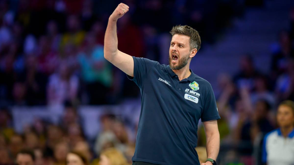 Trener piłki ręcznej w ciemnej koszulce z podniesioną prawą ręką, krzyczący z radości lub motywacji na tle trybun kibiców.