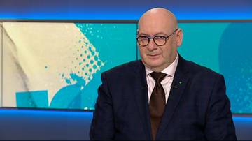 Mężczyzna w okularach i ciemnym garniturze, z brązowym krawatem, siedzi przed ekranem w studiu telewizyjnym. Ma łysą głowę i patrzy prosto przed siebie z lekkim uśmiechem. Tło za nim to abstrakcyjny wz&oacute;r w odcieniach niebieskiego i białego.