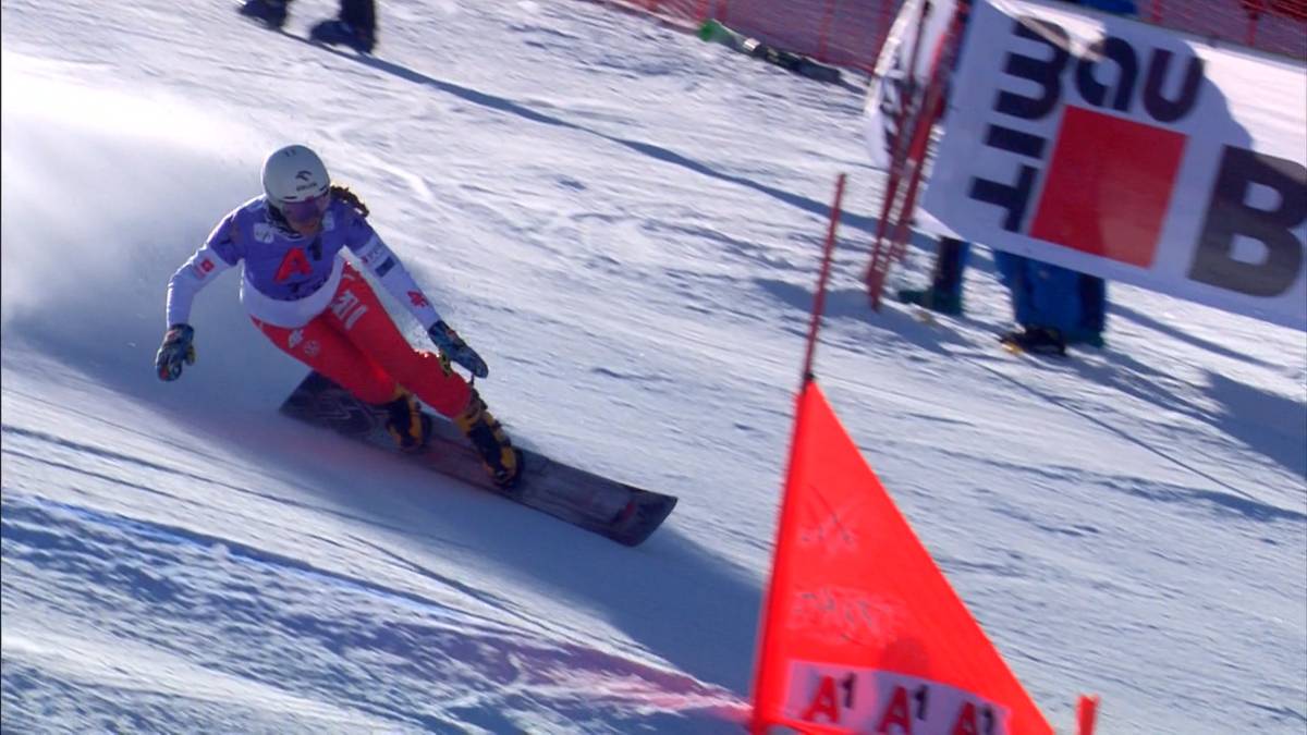 Snowboardzistka w czerwonych spodniach i białej kurtce z niebieskimi elementami zjeżdża po ośnieżonym stoku, mijając czerwoną tyczkę.