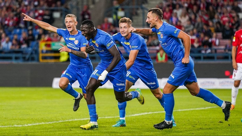 Czterech piłkarzy w niebieskich strojach cieszy się z bramki, jeden z nich podnosi rękę w geście zwycięstwa. Na drugim planie widoczni są kibice na trybunach.