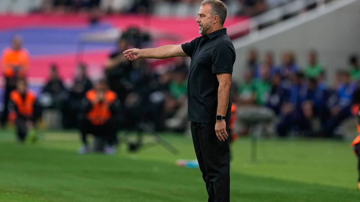 Trener piłkarski w czarnym polo i czarnych spodniach, stojący na boisku, wskazujący ręką w bok.