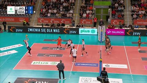 Drużyna JSW Jastrzębski Węgiel gra w meczu z Energą Trefl Gdańsk. Widoczni zawodnicy na boisku, sędzia oraz kibice na trybunach.