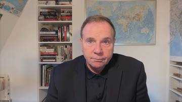 Mężczyzna w czarnym garniturze i czarnej koszuli siedzi przed półkami z książkami i mapą świata na ścianie.