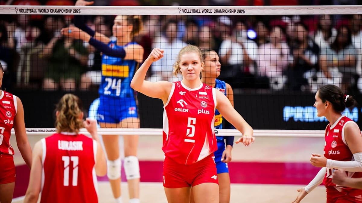 Siatkarka w czerwonej koszulce z numerem 5, uśmiecha się i unosi rękę. W tle inne zawodniczki i siatka.