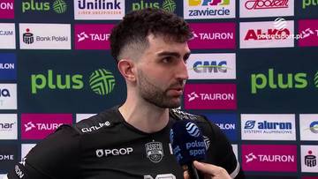 Mężczyzna w czarnej koszulce sportowej z mikrofonem przed twarzą, na tle sponsor&oacute;w.