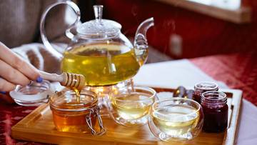 Szklany dzbanek z gorącym napojem, obok dwa kieliszki, słoiczek z miodem i drewniana chochla, osoba dodaje miód do napoju.