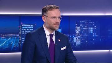 Mężczyzna w granatowym garniturze, białej koszuli i fioletowym krawacie z wąsami i okularami, na tle ekranów z nocnym widokiem miasta.