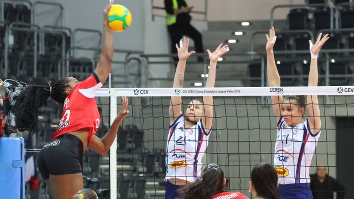 Siatkarka w czerwonej koszulce wykonuje atak nad siatką, podczas gdy dwie zawodniczki w białych koszulkach pr&oacute;bują ją zablokować.