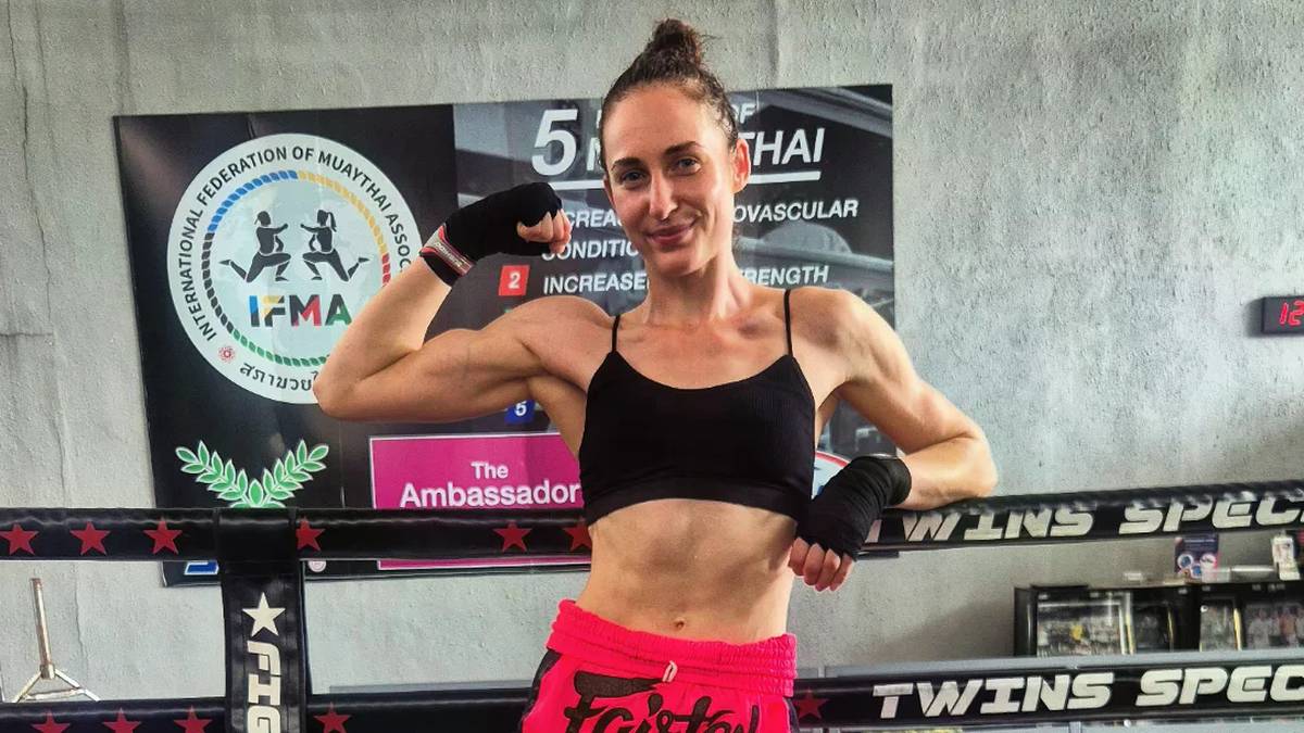 Kobieta w stroju sportowym, z rękami opartymi o linę ringu, demonstruje biceps. W tle widoczne jest logo federacji Muay Thai.