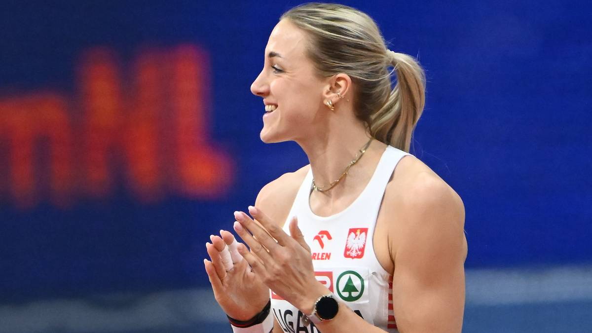 Atletka w białej koszulce sportowej z białym i czerwonym logo Polski, z uśmiechem na twarzy, klaszcze w dłonie. Jej jasne włosy są upięte w kucyk.