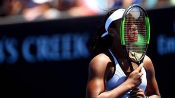 Tenisistka trzymająca rakietę przed twarzą podczas meczu, z rozmytym tłem sponsor&oacute;w.