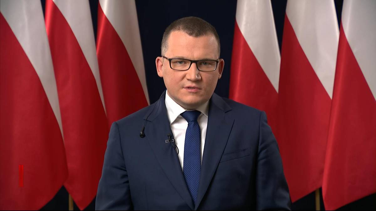 Mężczyzna w granatowym garniturze i niebieskim krawacie, noszący okulary, stoi przed rzędem polskich flag. Patrzy prosto przed siebie z poważnym wyrazem twarzy.