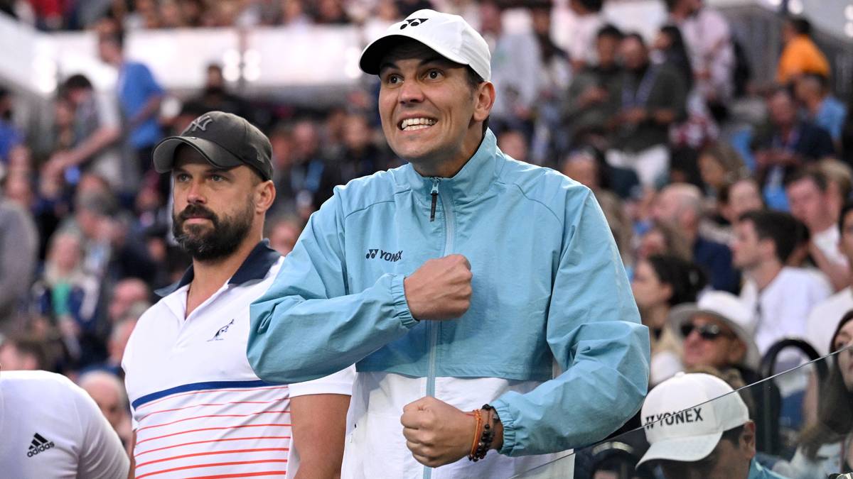 Dwaj mężczyźni w strojach sportowych na tle publiczności podczas zawodów tenisowych. Mężczyzna na pierwszym planie w jasnoniebieskiej kurtce sportowej z białą wstawką, ma na głowie białą czapkę z daszkiem i zaciśnięte pięści w geście radości. Obok niego stoi mężczyzna w czapce z daszkiem i koszulce z pasami.