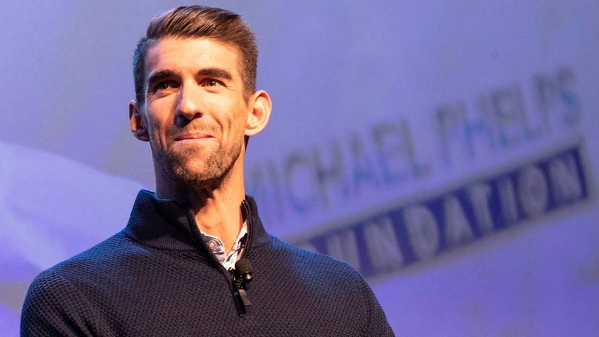 Michael Phelps uśmiecha się, ubrany w ciemny sweter. W tle widoczne jest logo "Michael Phelps Foundation".
