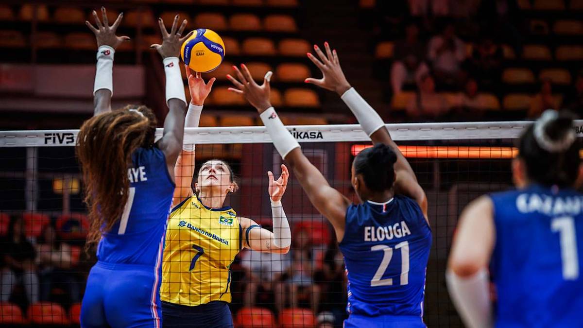 Trzy siatkarki w akcji przy siatce, jedna z nich z piłką, próbuje ją odbić. Dwie pozostałe starają się zablokować atak.