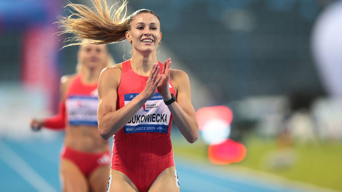 Lekkoatletka Natalia Kaczmarek w czerwonym stroju sportowym z biało-niebieskim pasem na piersi, z uśmiechem na twarzy i splecionymi dłońmi, podczas biegu na stadionie.