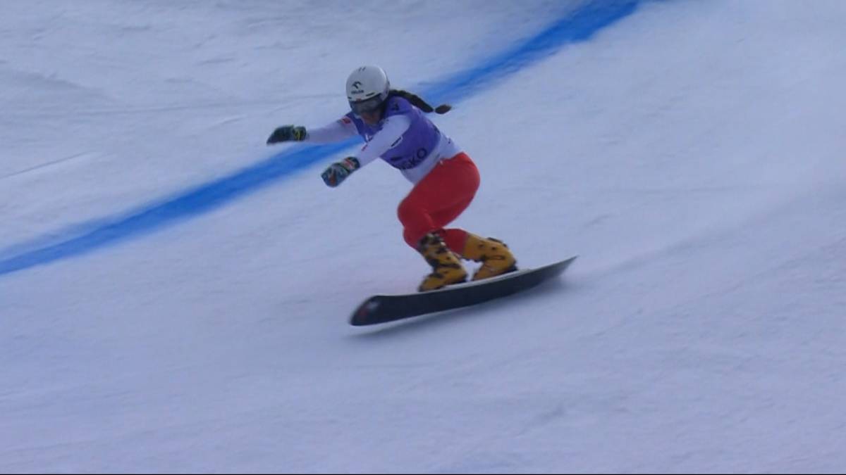 Snowboarderka w czerwonych spodniach i fioletowej bluzie jedzie po ośnieżonym stoku, pokonując niebieską trasę wyznaczoną przez tyczki.