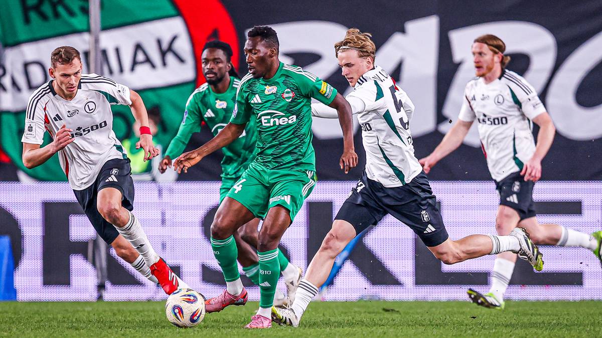 Piłkarze w białych i zielonych strojach rywalizują o piłkę na boisku piłkarskim.