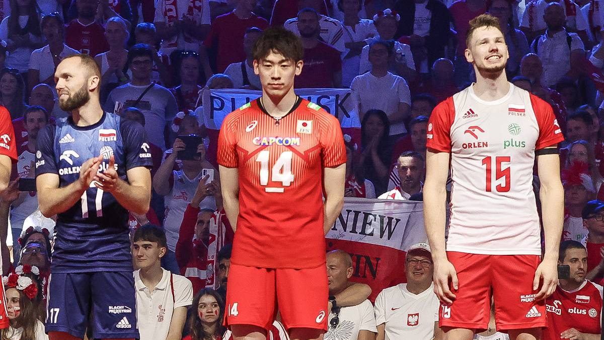 Trzech siatkarzy, jeden w niebieskiej koszulce z numerem 11, drugi w czerwonej koszulce z numerem 14, trzeci w białej koszulce z numerem 15, stoi przed publicznością.