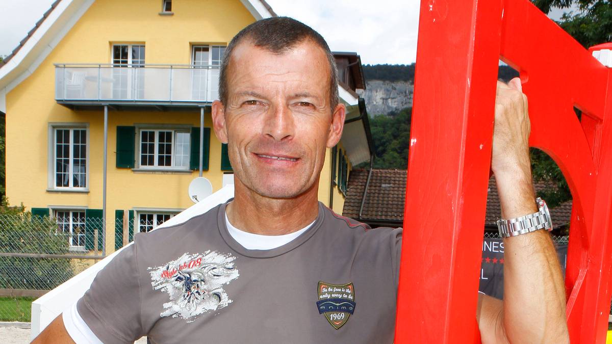 Portret mężczyzny w średnim wieku, uśmiechniętego, w szarym t-shircie, stojącego obok czerwonej konstrukcji, na tle ż&oacute;łtego domu.