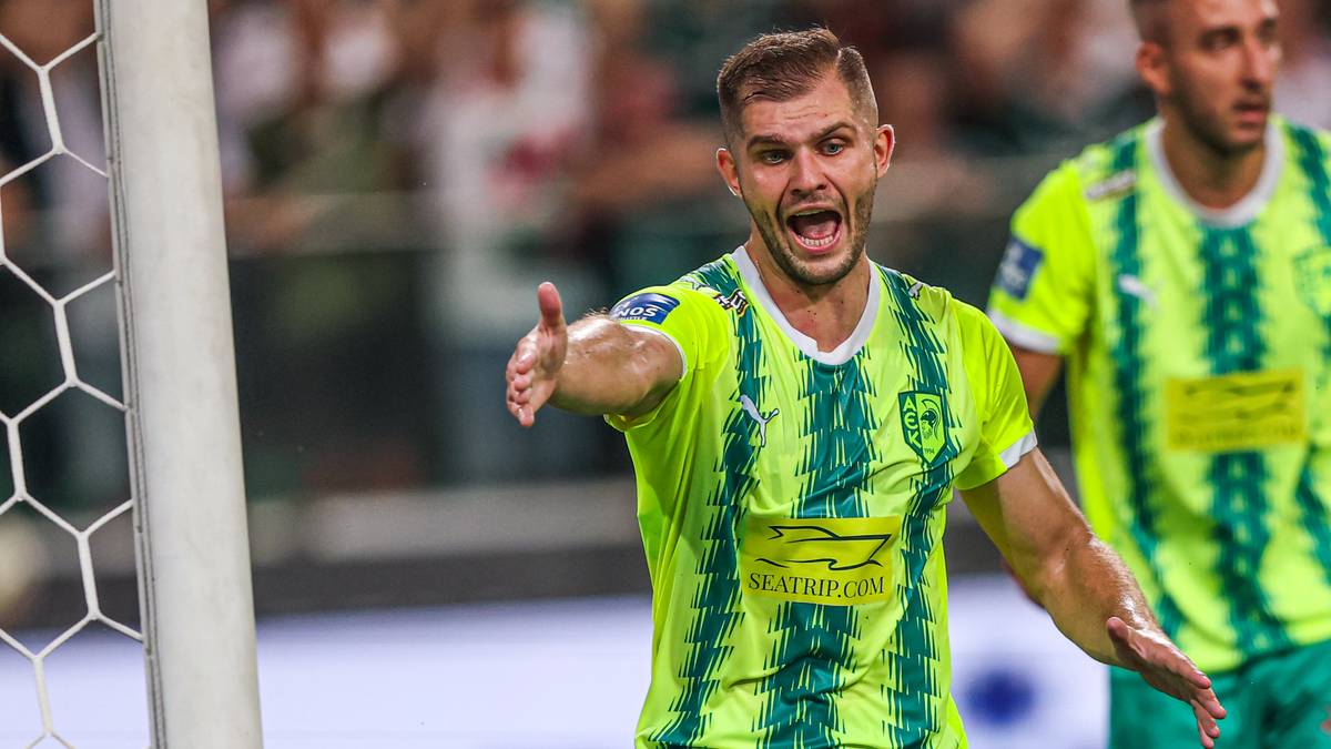 Piłkarz w zielono-ż&oacute;łtej koszulce z wyciągniętą ręką i otwartymi ustami na boisku.