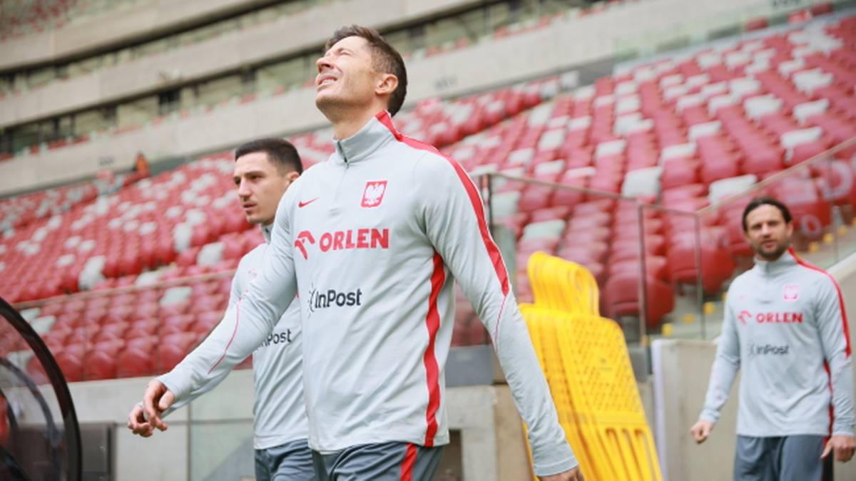 Robert Lewandowski w szarym dresie reprezentacji Polski z widocznym logo "ORLEN" patrzy w g&oacute;rę, na tle trybun stadionu.