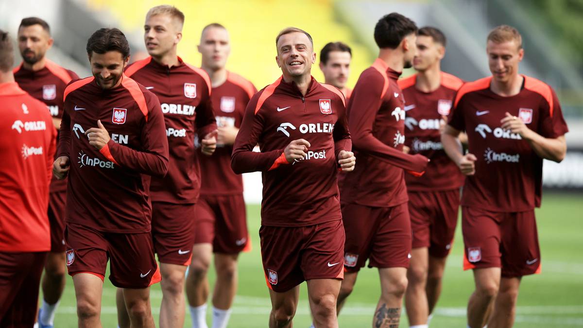 Grupa piłkarzy w bordowych strojach treningowych biegnie na zielonej trawie boiska.