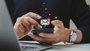 Dłoń osoby trzymającej smartfon, na ekranie którego wyświetlona jest ikona e-mail z powiadomieniem. W tle widać fragment laptopa.