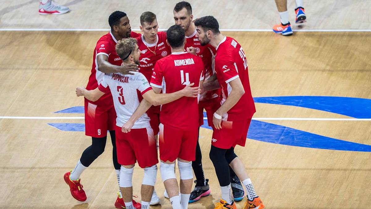 Siedmiu siatkarzy w czerwonych strojach sportowych stoi w okręgu na boisku siatkarskim, spoglądając na siebie i okazując radość i wsparcie.