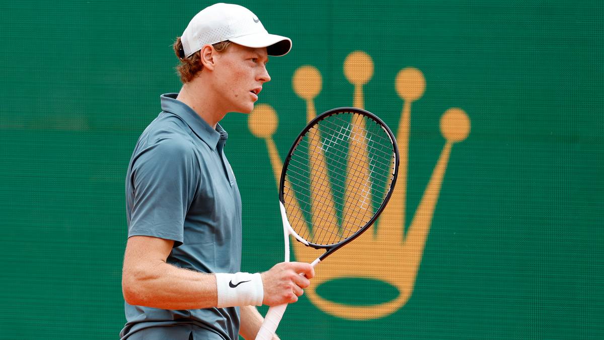 Młody tenisista w białej czapce i szarym polo, trzymający rakietę tenisową, na zielonym tle z ż&oacute;łtym logo Rolex.