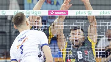 Siatkarz w białej koszulce odwrócony plecami, drugi zawodnik w zielono-żółtej koszulce blokuje piłkę nad siatką.