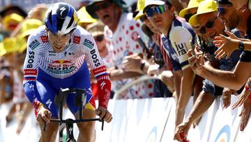 Kolarz w białym, niebieskim i czerwonym stroju z napisem "BOSS" jedzie na rowerze, podczas gdy widzowie w ż&oacute;łtych kapeluszach dopingują go zza barierki.