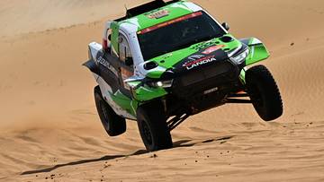 Zielono-biały samoch&oacute;d rajdowy jadący po piaszczystym terenie.