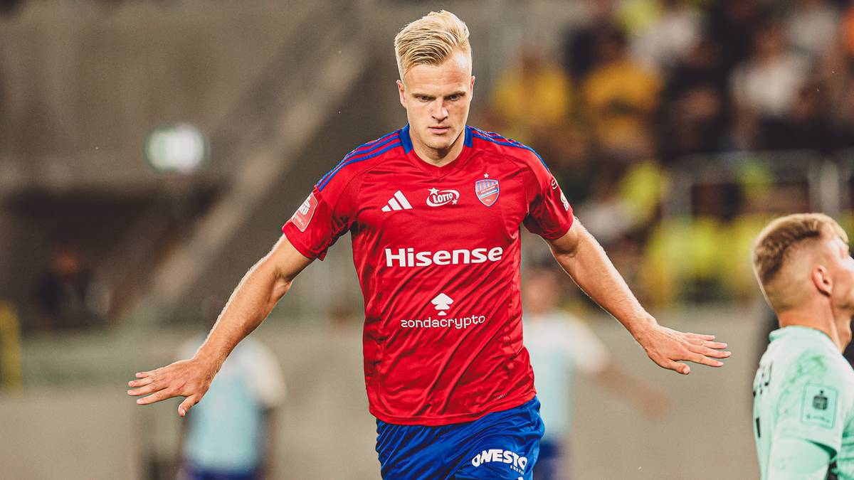 Piłkarz w czerwonej koszulce z herbem klubu Raków Częstochowa, stojący na boisku.