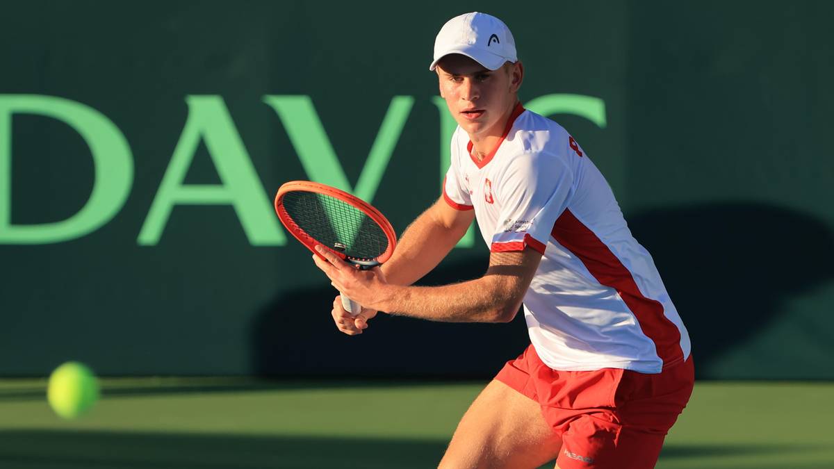 Tenisista w białej czapce i stroju sportowym uderza piłkę rakietą.