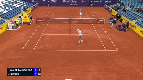Dwaj tenisiści grający mecz na korcie ziemnym. Jeden z nich, Botic Van de Zandschulp, jest bliżej kamery, a drugi, Marc-Andrea Huesler, znajduje się po drugiej stronie siatki. Widoczni są widzowie na trybunach.