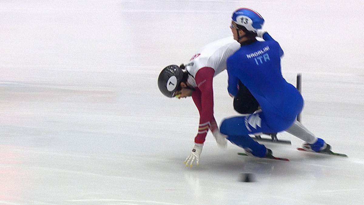 Dwóch zawodników short track na lodowisku, jeden upada, drugi stara się go wyprzedzić.