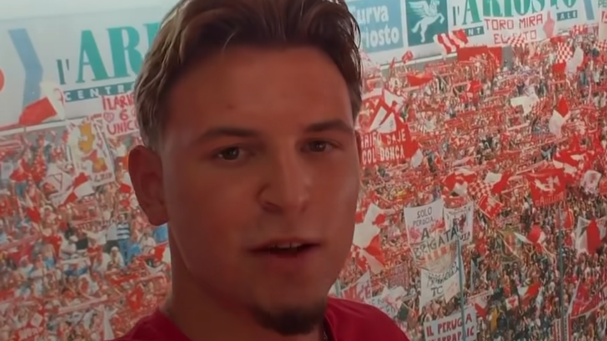 Młody mężczyzna z jasnymi włosami, ubrany w czerwoną koszulkę, pozuje do zdjęcia na tle ściany ozdobionej grafikami z motywami piłkarskimi, w tym licznymi flagami i kibicami.