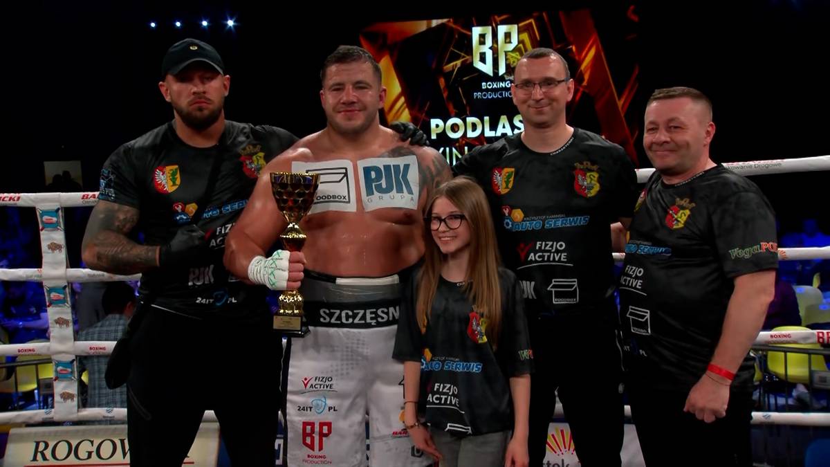 Pięciu mężczyzn i jedna dziewczynka stoją na ringu bokserskim, trzymając puchary i pozując do zdjęcia. Dw&oacute;ch mężczyzn po bokach ma na sobie czarne koszulki z logotypami, a mężczyzna pośrodku ma na piersiach napis "PJK". Dziewczynka stoi przed nim, a po jej lewej stronie znajduje się mężczyzna w okularach.