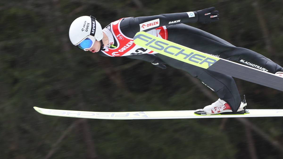 Skoczek narciarski w powietrzu, pochylony do przodu, na tle drzew.