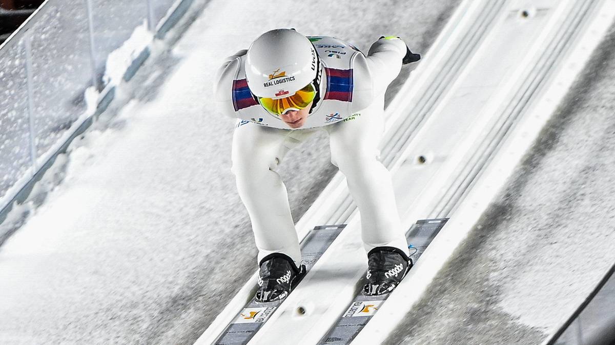 Skoczek narciarski w białym stroju z goglami na nosie zjeżdża po rozbiegu skoczni narciarskiej.